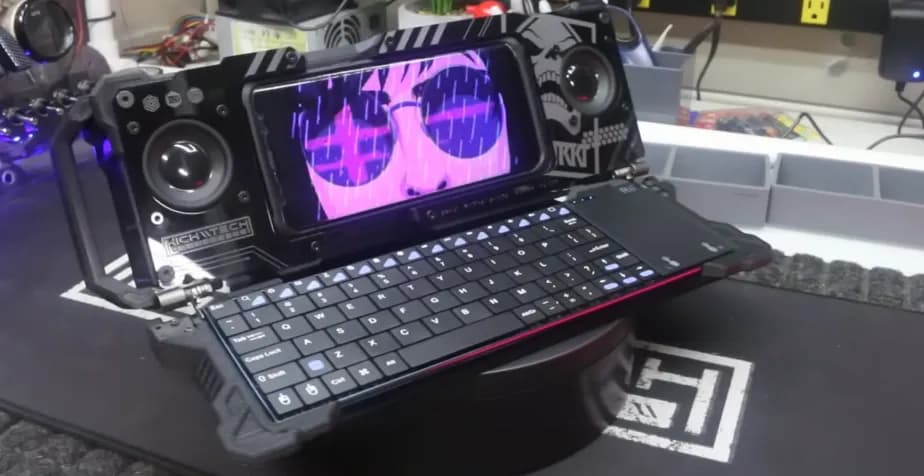 Cyberdeck clamshell berwarna gelap dalam posisi terbuka, menampilkan keyboard kompak dan area untuk meletakkan ponsel, dengan desain retro-futuristik.