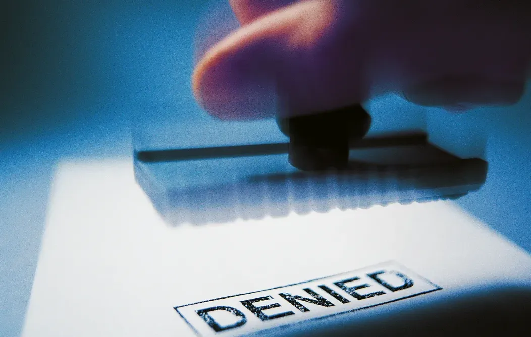 Stempel bertuliskan "DENIED" atau "DITOLAK" ditekan di atas dokumen, simbol penolakan klaim asuransi oleh sistem otomatis.