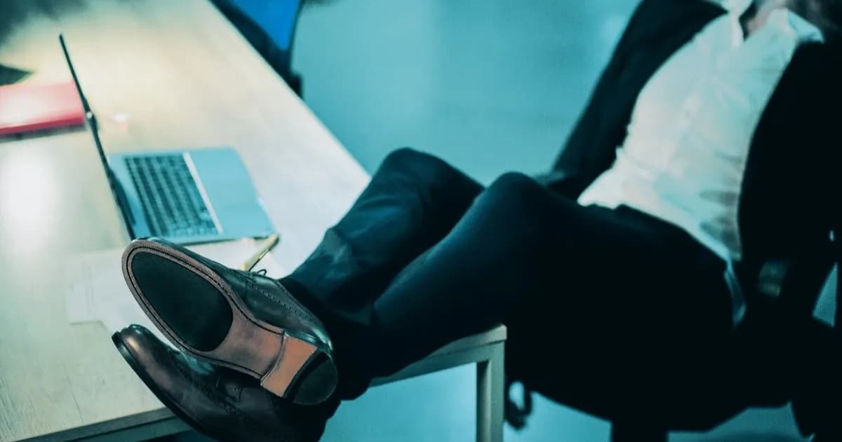 Ilustrasi seorang pekerja duduk santai dengan kaki di atas meja kantor, di samping laptop yang sedang digunakan