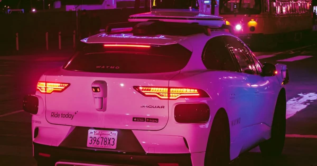 Mobil listrik Jaguar putih dengan sensor otonom Waymo di atap dan belakang, diparkir di jalan kota pada malam hari dengan lampu belakang menyala dan plat nomor California. Mobil memiliki stiker 'Ride today' dan app store di bagian belakang. Trem terlihat di latar belakang.