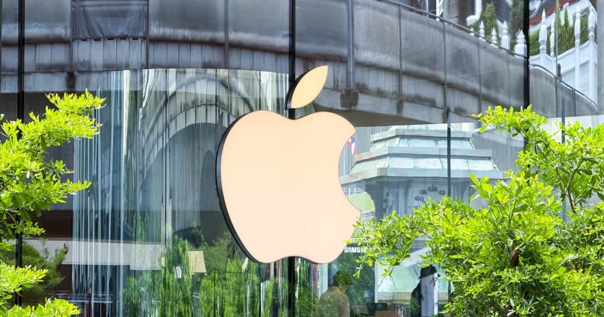 Gedung kaca dengan logo Apple di eksterior, dua pohon hijau terlihat dan terpantul di struktur bangunan