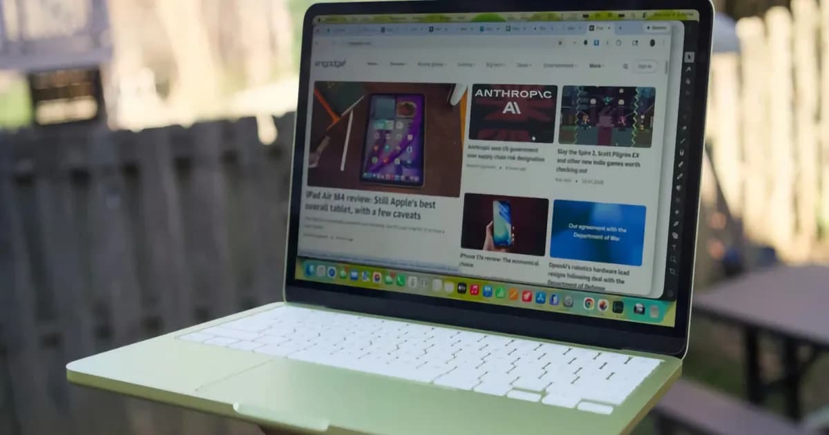 Tangan memegang MacBook Neo warna kuning yang terbuka di atas meja kayu.