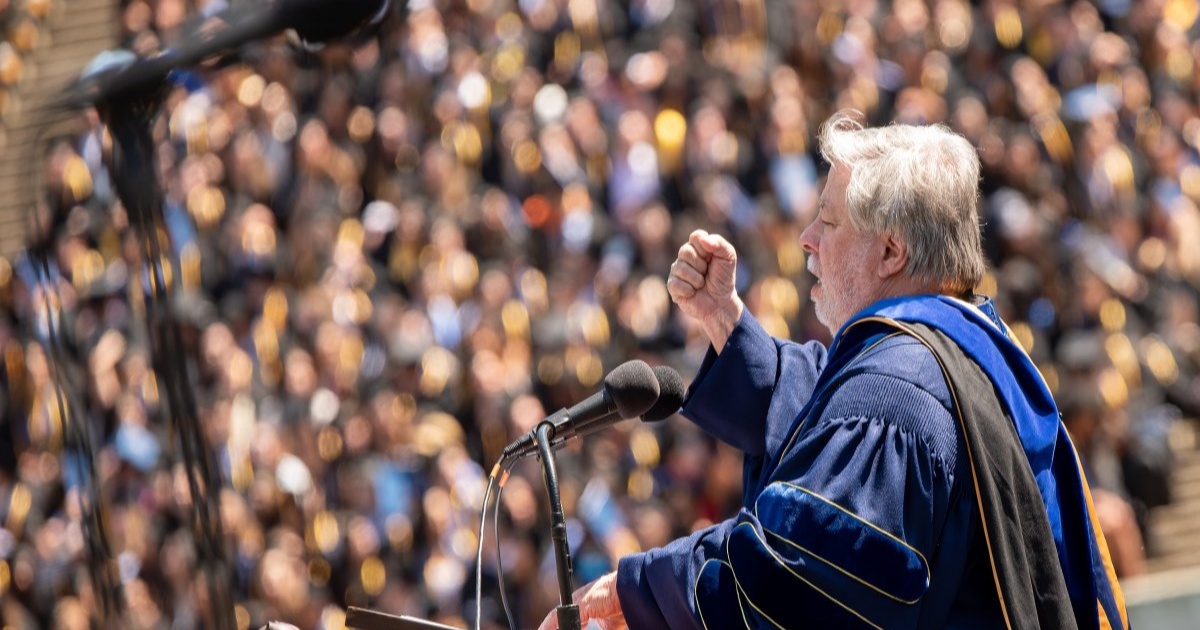 Steve Wozniak berpidato di podium wisuda dengan gestur tangan terkepal, mengenakan jubah akademik biru