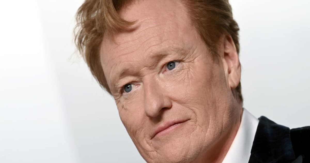 Conan O’Brien tersenyum di depan latar putih mengenakan jas