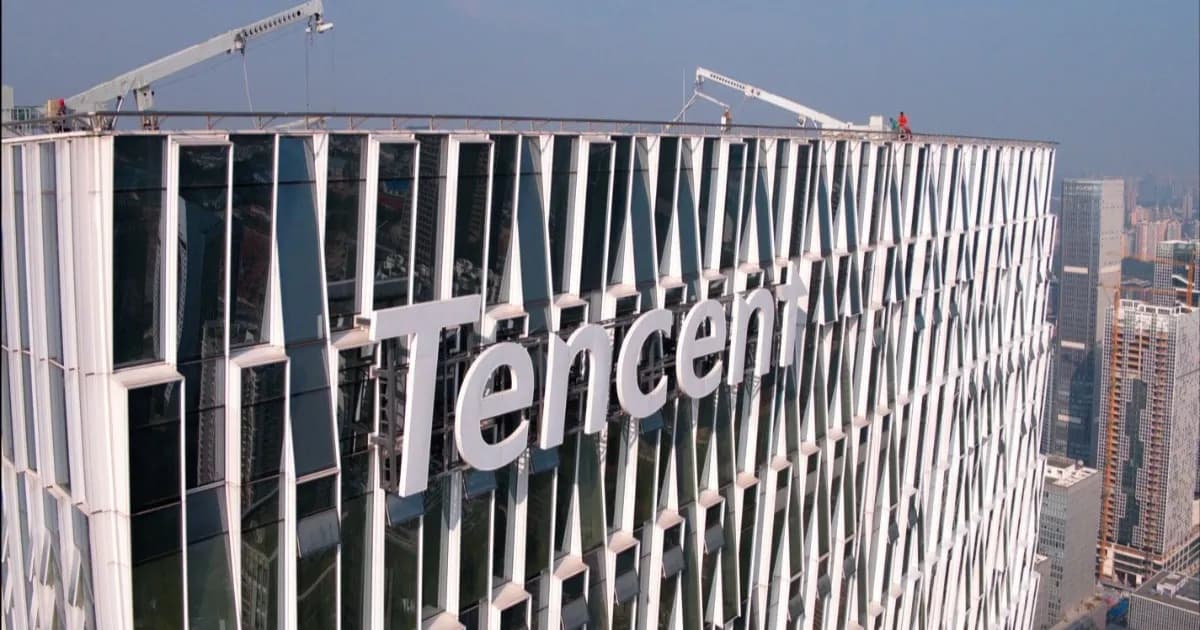 Logo Tencent berwarna putih besar pada fasad menara kantor kaca dengan crane atap dan skyline kota di belakangnya