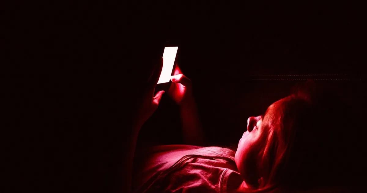 Remaja perempuan berbaring di sofa dalam kondisi gelap sambil menggunakan smartphone.